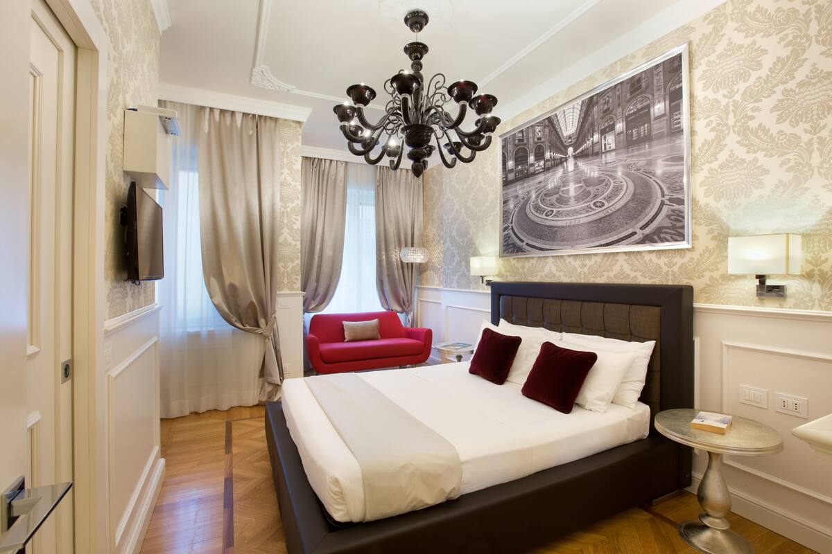 A hotel room with a large bed, a red armchair, a chandelier, a flat-screen TV, and a wall-mounted artwork.