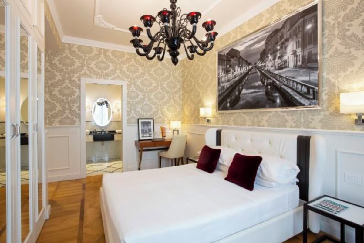 A hotel room with a large bed, a chandelier, a desk with a chair, a mirror, and a framed picture on the wall.