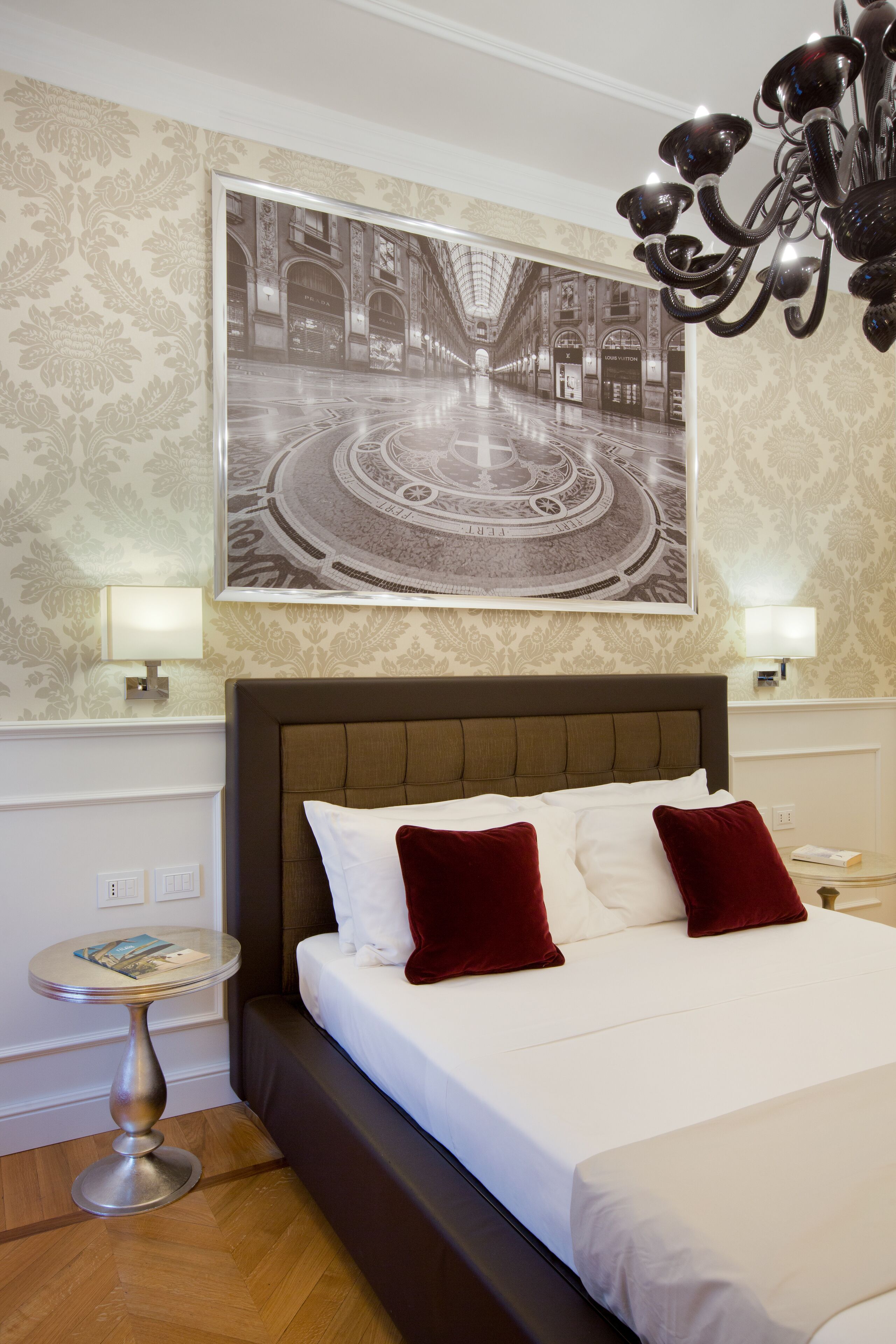Hotelkamer met een groot bed, twee rode kussens, een rond bijzettafeltje, een aan de muur hangend schilderij en een kroonluchter.