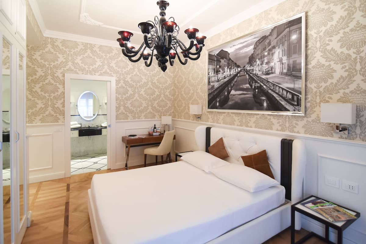 A hotel room with a bed, a desk, a chair, a chandelier, a wall-mounted mirror, and a framed picture.