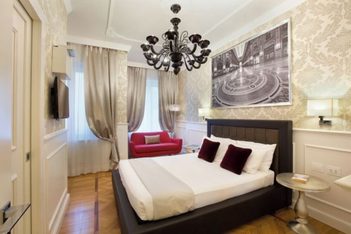 A hotel room with a large bed, a red sofa, a chandelier, a painting, and a television.