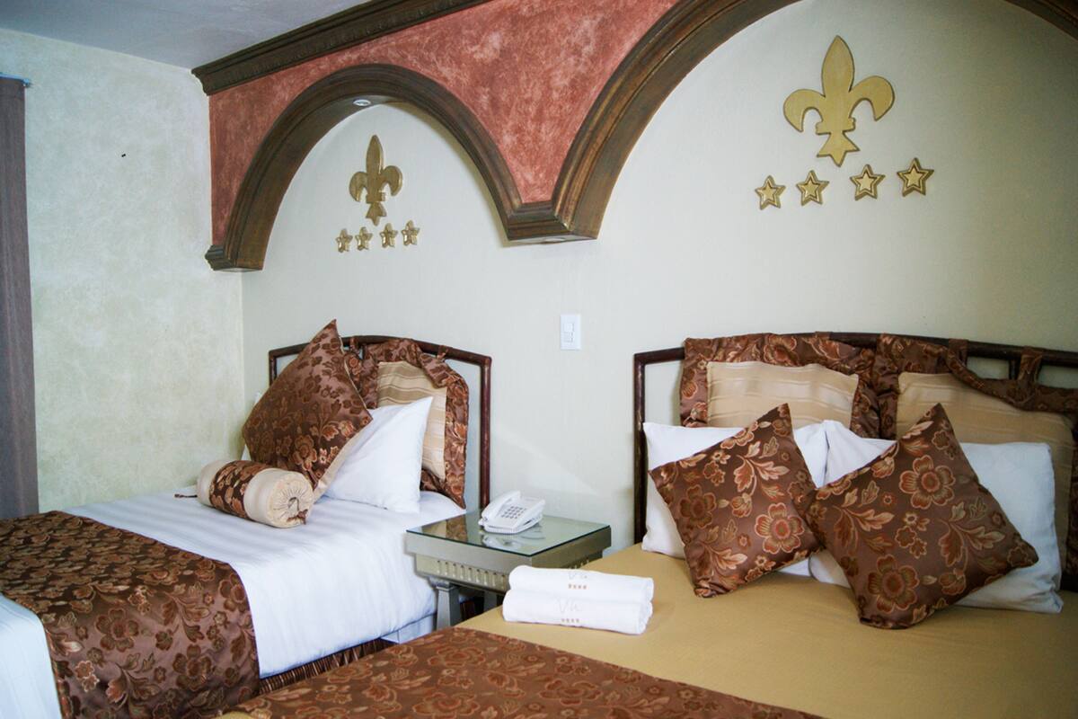 A hotel room with two beds, each with floral-patterned bedding and pillows, a bedside table with a phone, and a wall with fleur-de-lis and star decorations.