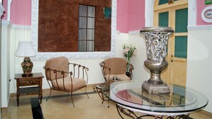 A room with a pink wall, a large window with a brown curtain, a round glass table, two chairs, a small table with a lamp, and a decorative vase. - Villahernan Hotel (Puebla)