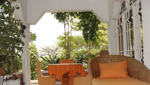 Un espacio exterior cubierto con un sofá de mimbre y una mesa, rodeado de plantas en macetas y vistas a árboles.