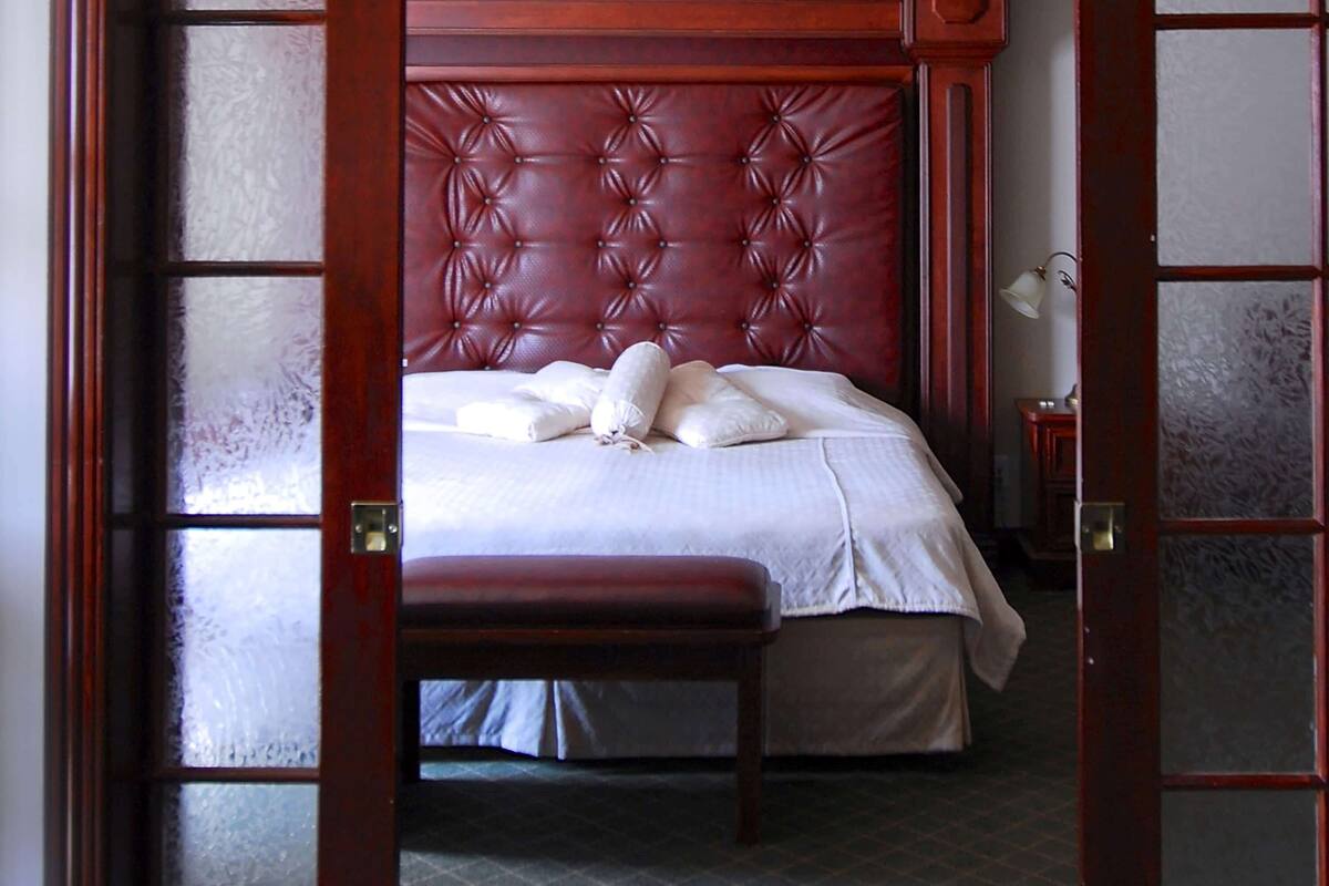 Une chambre à coucher avec une tête de lit capitonnée, un lit avec du linge de lit blanc, un banc et une table de chevet avec une lampe.