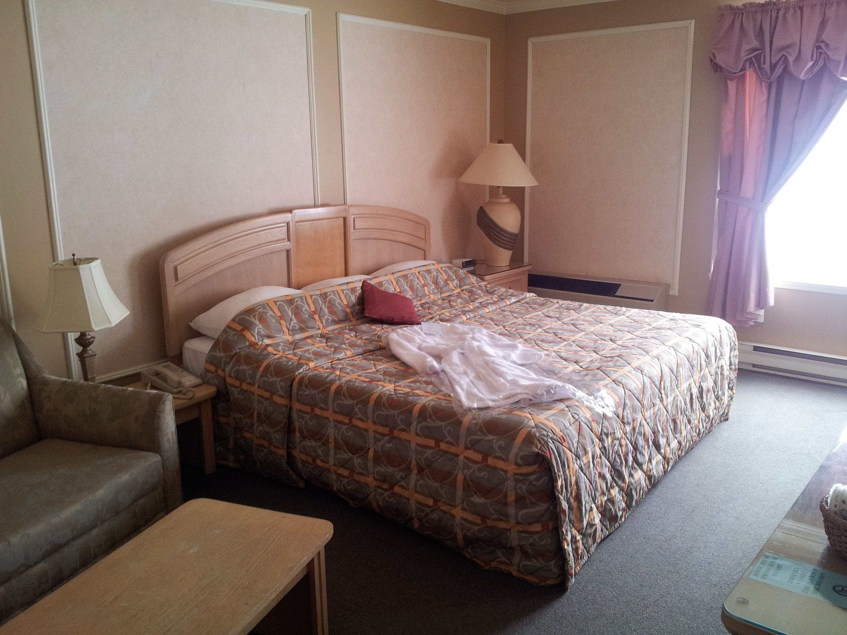 Una habitación de hotel con cama, una silla, una mesita de noche, una lámpara y una ventana con cortinas.
