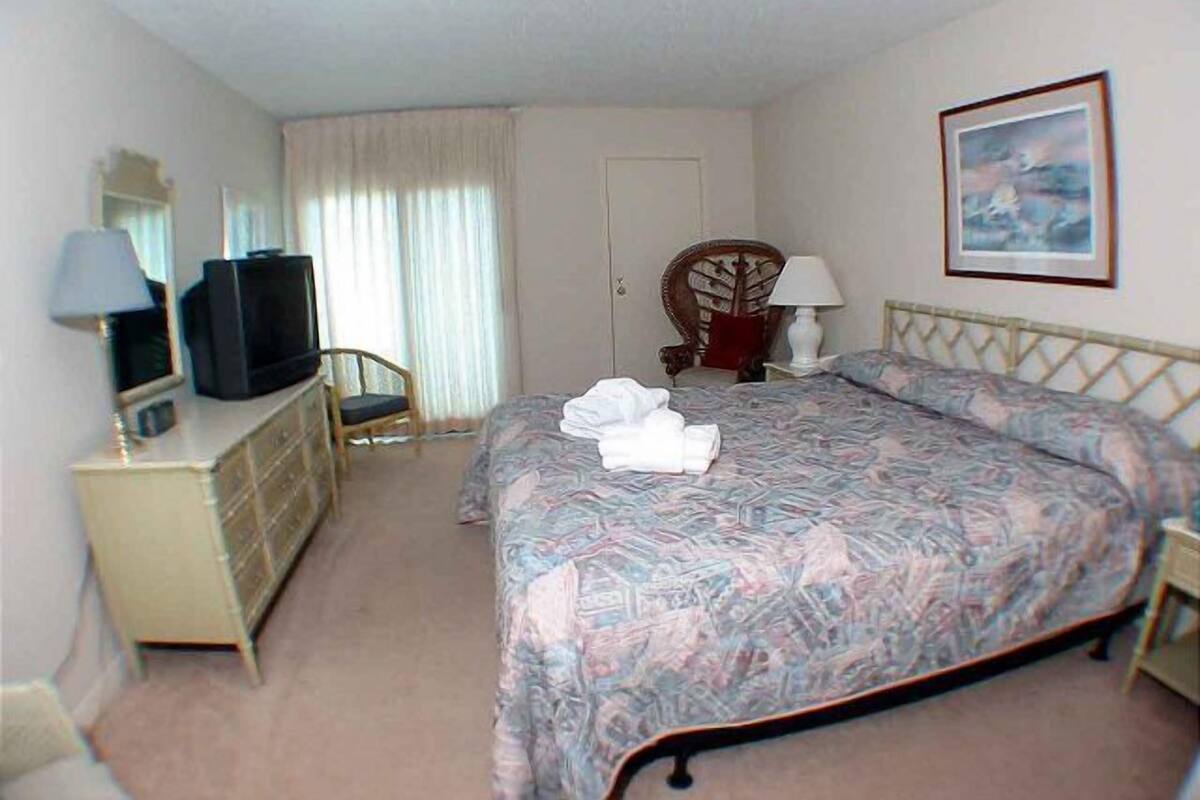 Habitación de hotel con cama, televisor, cómoda, silla y un cuadro en la pared.