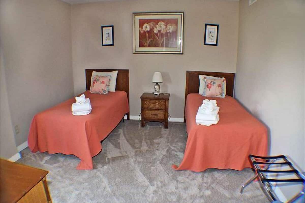 Habitación de hotel con dos camas, cada una con colcha y almohadas rojas, una mesita de noche de madera con lámpara, y un cuadro en la pared.