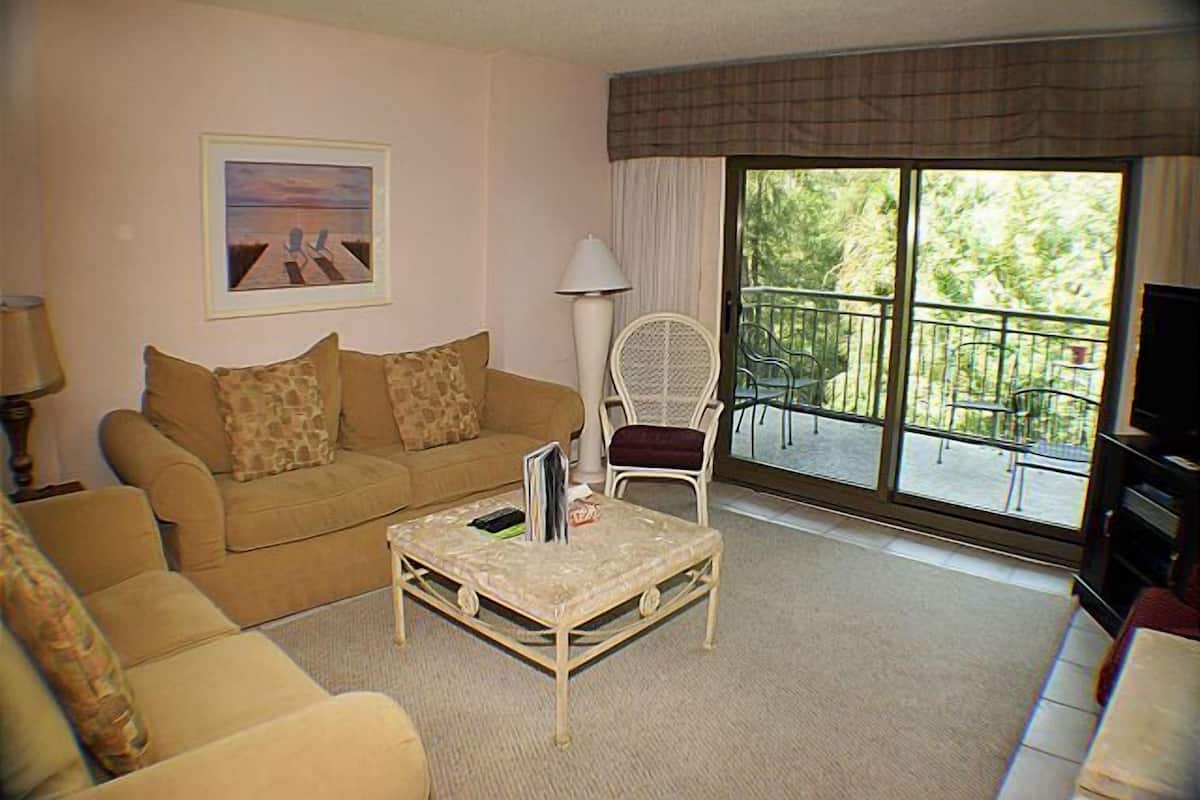 Una sala de estar con un sofá beige, un sillón blanco, una mesita y un balcón con mobiliario exterior.