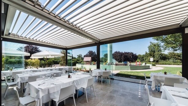 Un espace de restauration en plein air avec des tables et des chaises, une structure vitrée et une vue sur un parc.