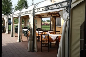 Un área de comedor al aire libre con mesas y sillas bajo una estructura cubierta con detalles de metal decorativo.