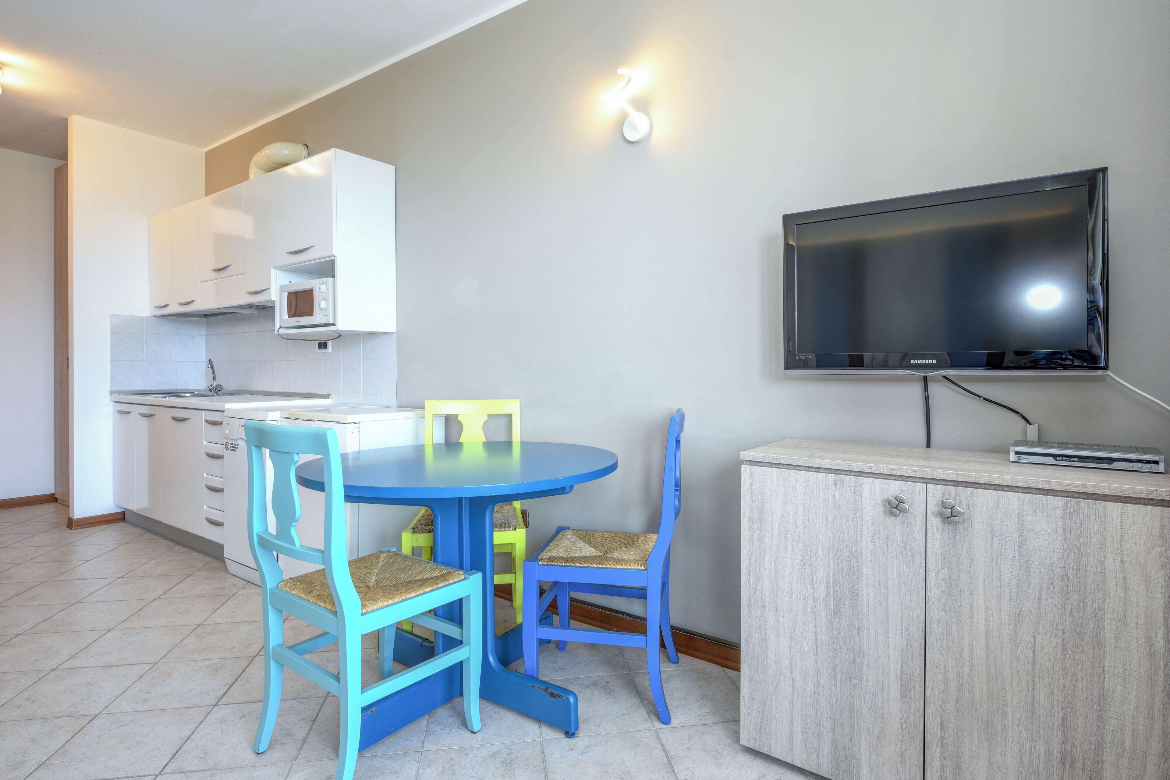 Una pequeña cocina con armarios blancos, un microondas y una mesa azul con dos sillas.