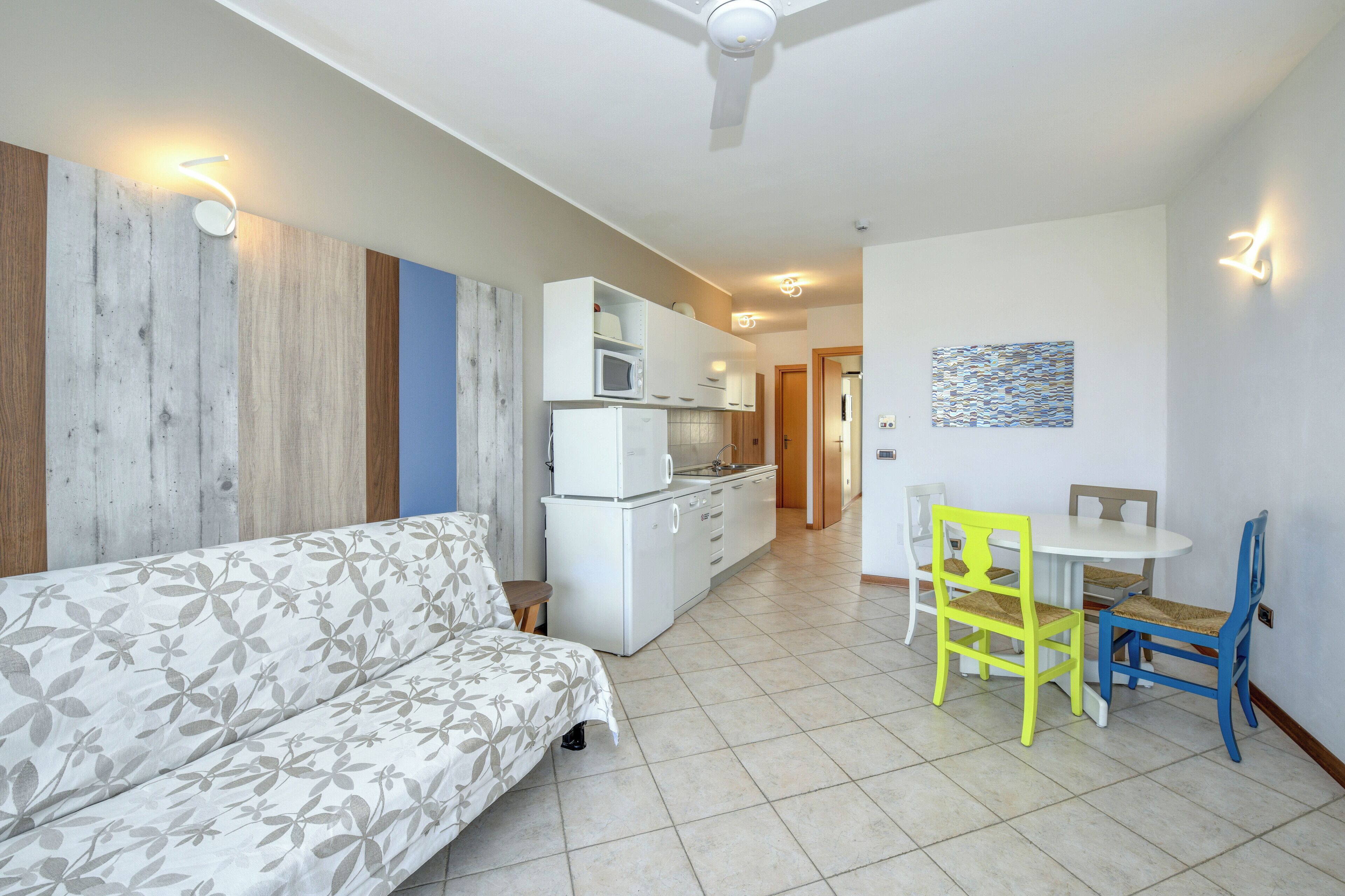 A modern living room with a sofa, a dining table with chairs, a kitchen area with cabinets and appliances, and a ceiling fan.