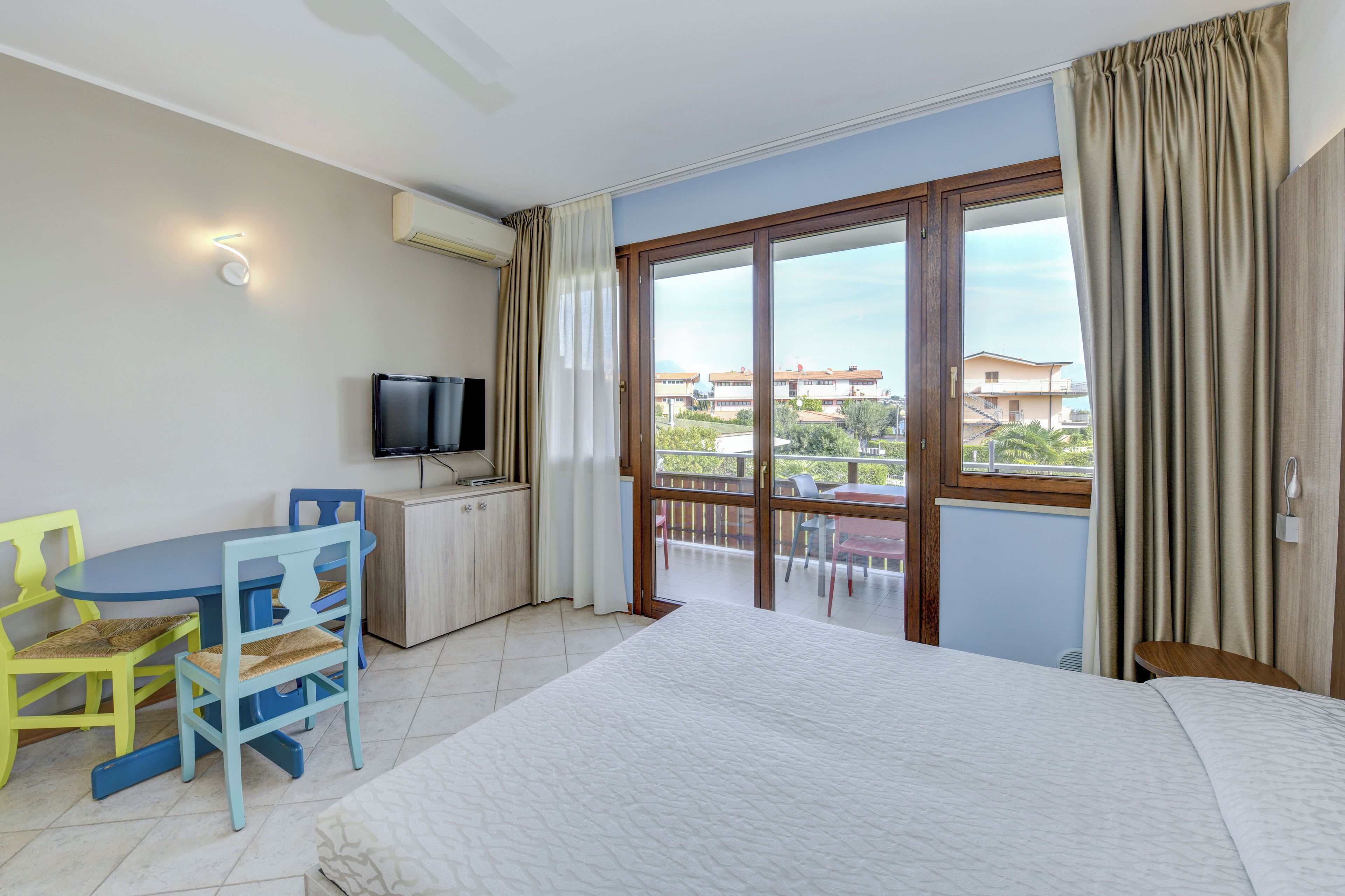 Une chambre d’hôtel avec un lit, un bureau avec une télévision, un balcon donnant sur les bâtiments et des rideaux.