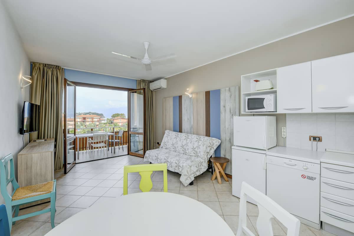A modern hotel room with a balcony, a sofa, a dining table, and a kitchenette.