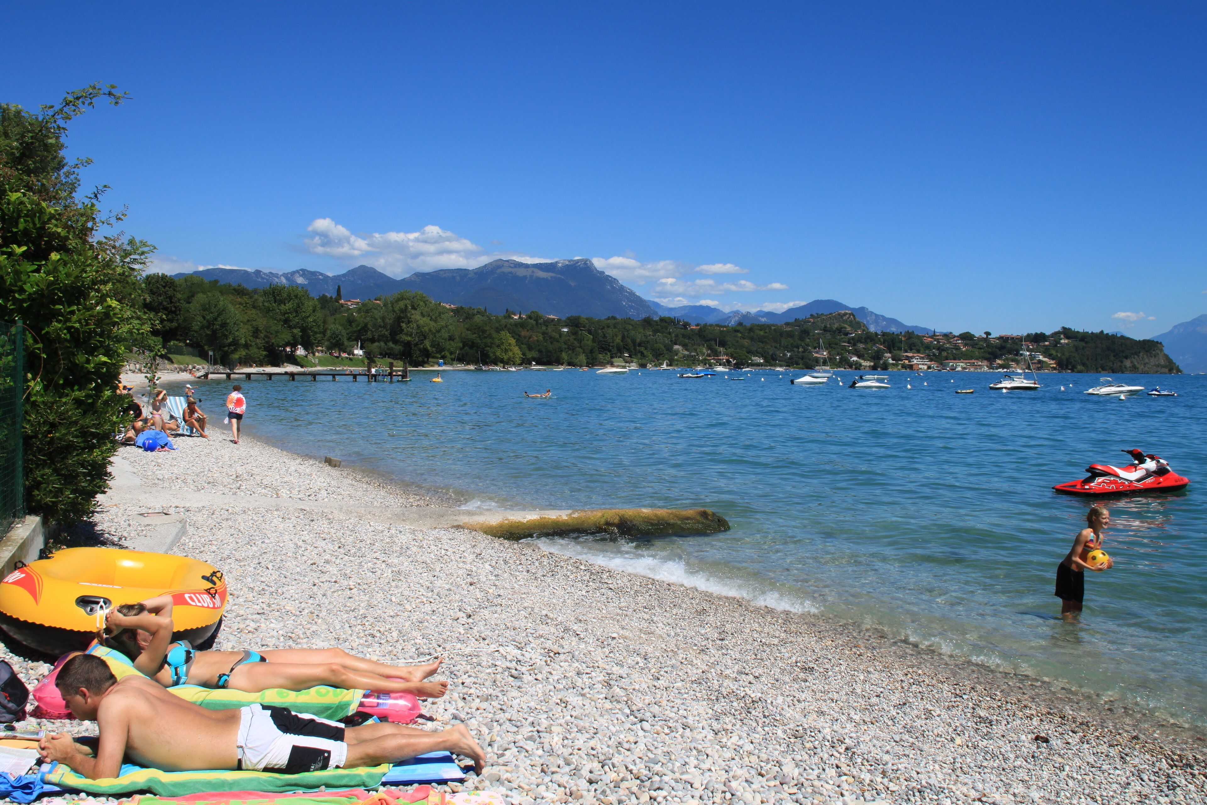 Una playa de guijarros con gente tomando el sol y nadando, barcos en el agua y montañas al fondo.