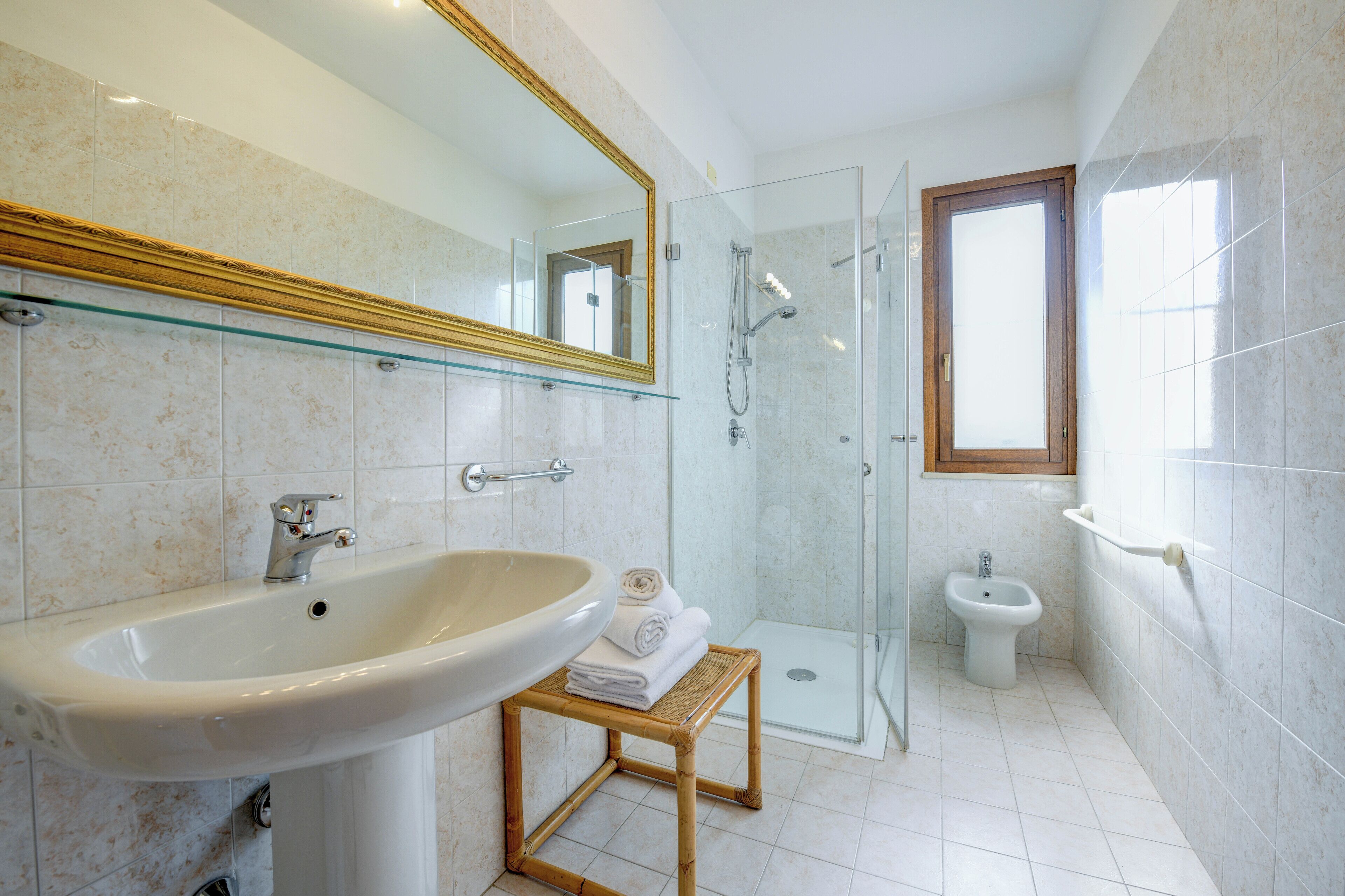 A modern bathroom with a large sink, a glass shower enclosure, a bidet, and a small stool with towels.