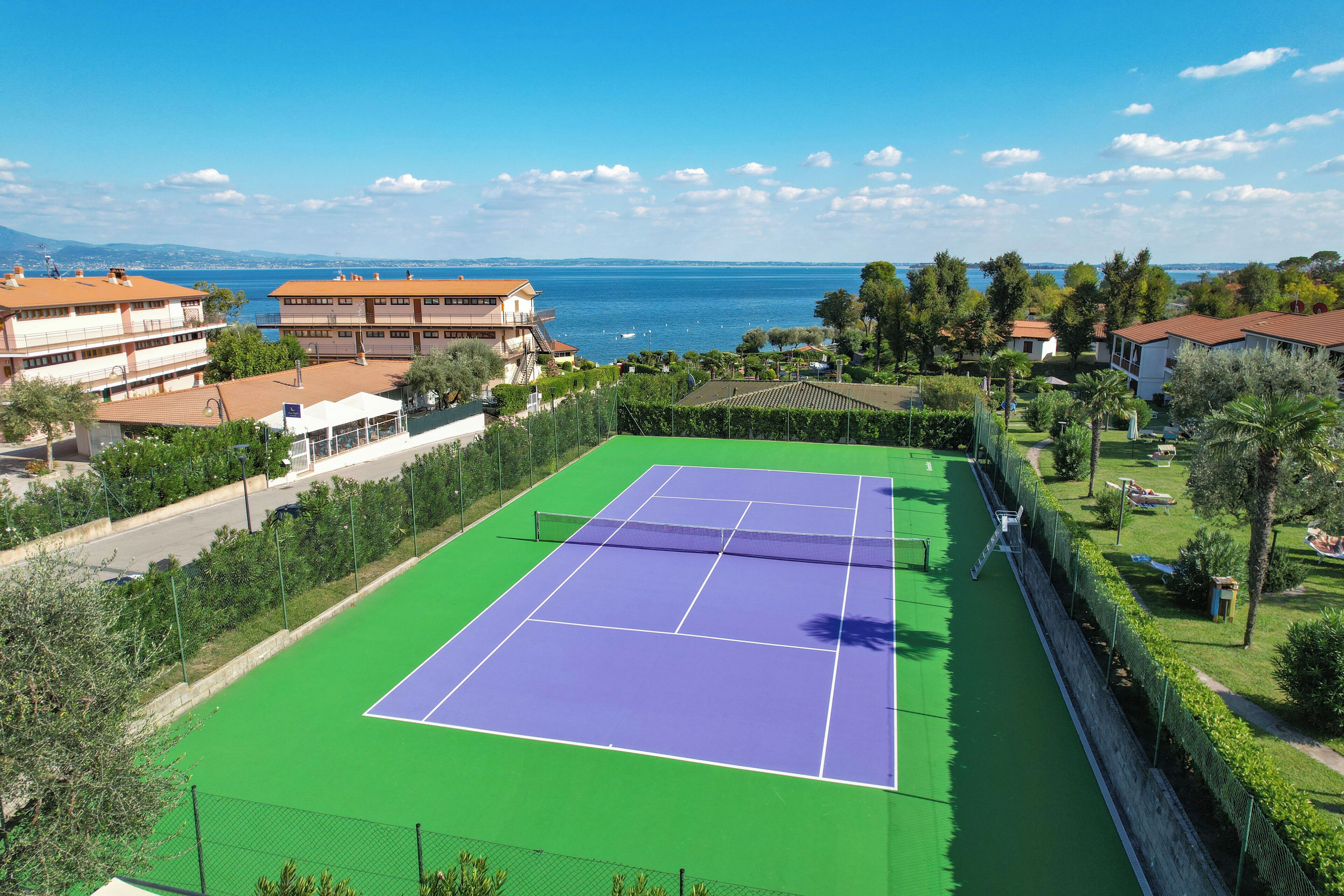 Un court de tennis avec un filet, entouré de verdure et de bâtiments, avec une vue sur la mer en arrière-plan.