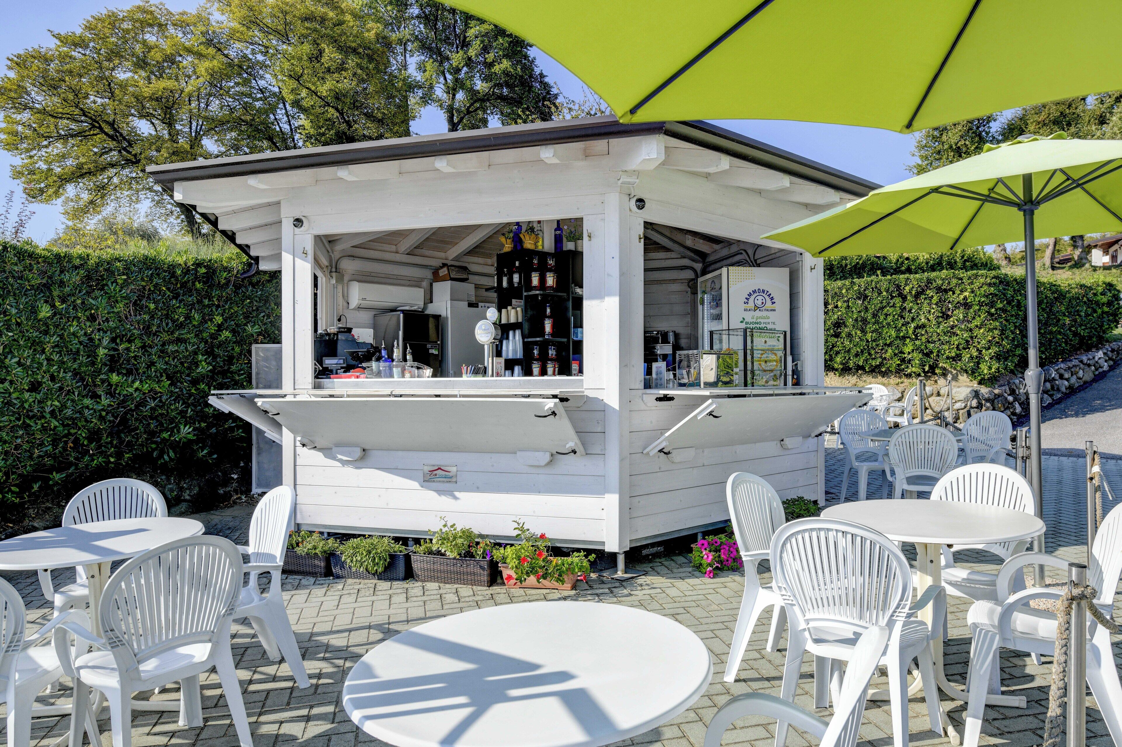 A white outdoor café with tables and chairs, a green umbrella, and a small building with shelves of bottles and a sign.