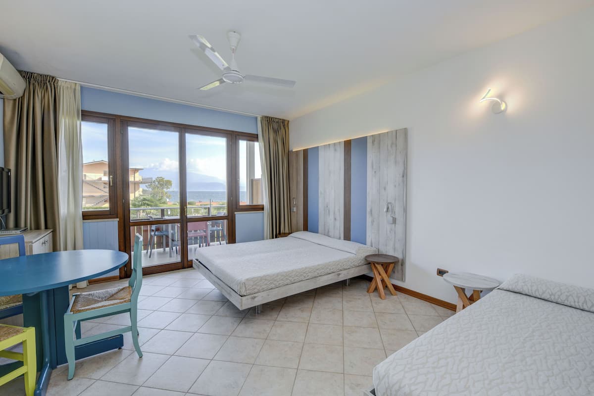 A hotel room with a bed, a desk, a chair, a small table, a ceiling fan, and a large window with a view of the sea.