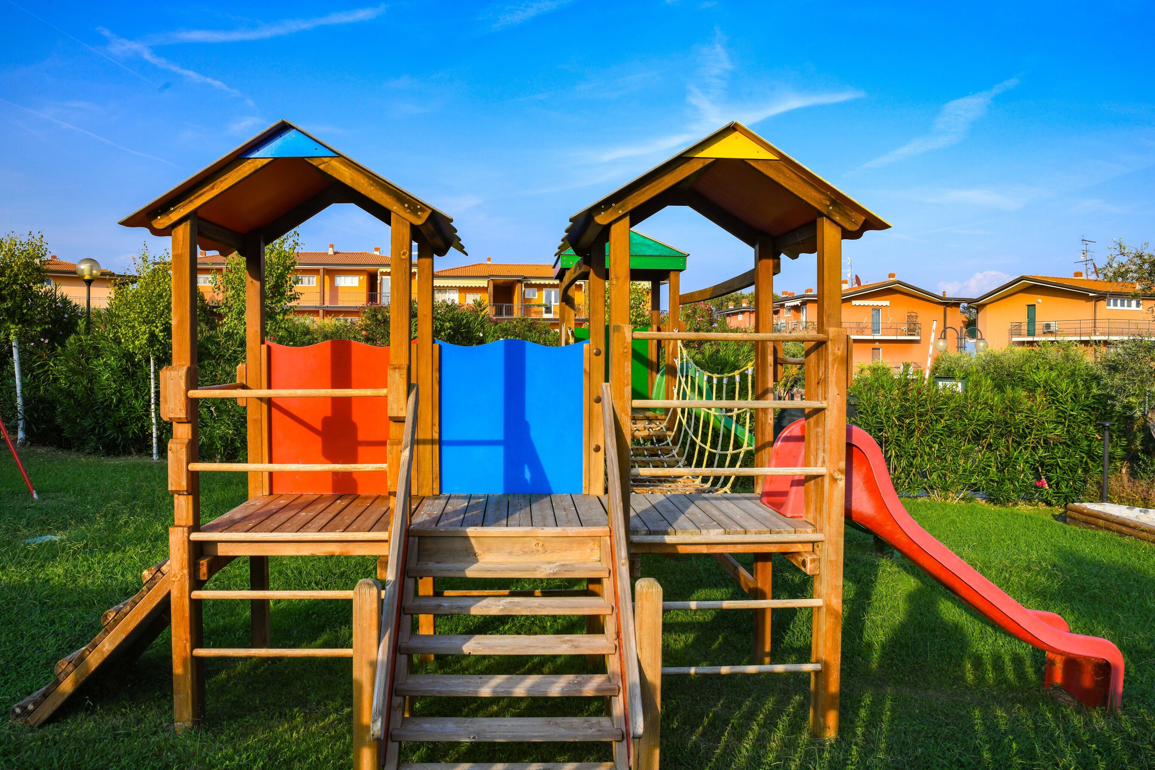 Estructura de juego de madera con tobogán y escaleras de acceso, rodeada de vegetación y edificios residenciales.