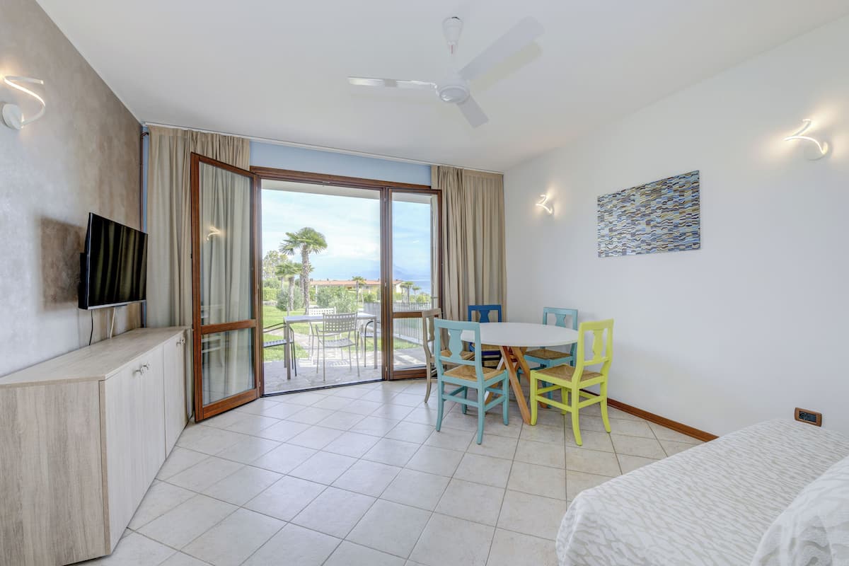 A modern hotel room with a balcony, a flat-screen TV, a dining table with chairs, and a bed.