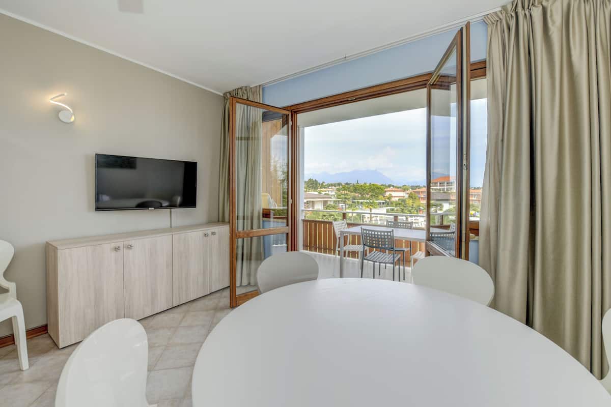 A modern room with a large window, a flat-screen TV, a dining table, and a view of a cityscape.