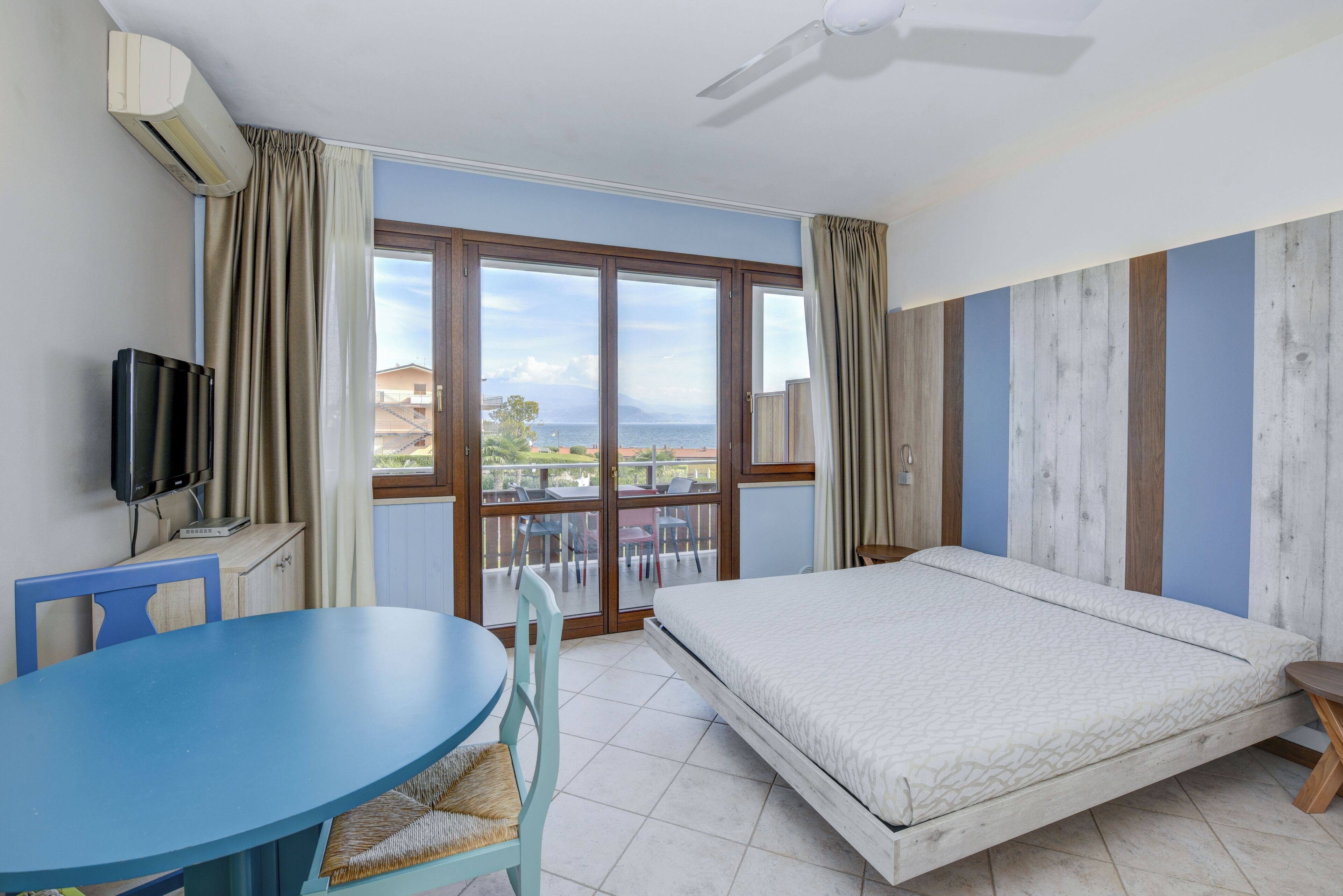 A hotel room with a bed, a TV, a table, and a view of the sea through large windows.