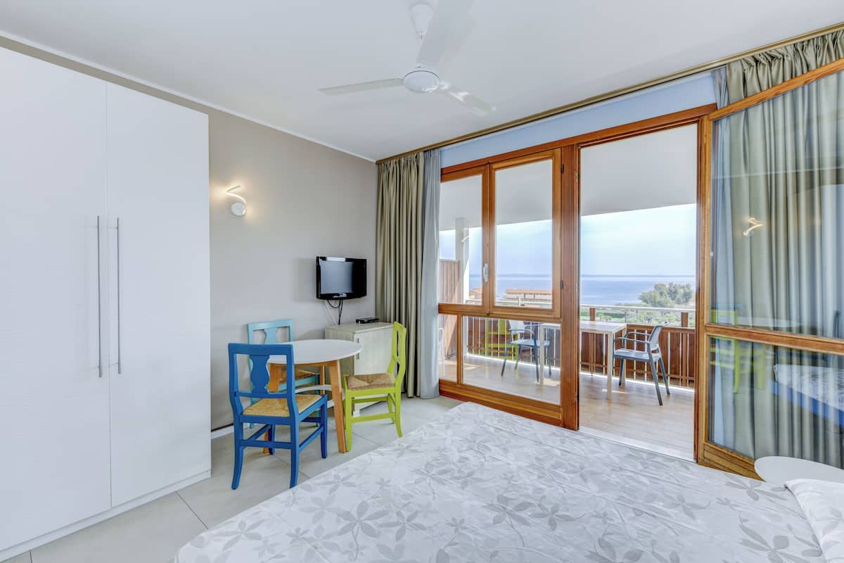 A bedroom with a bed, a desk, two chairs, a television, and a balcony with a view.