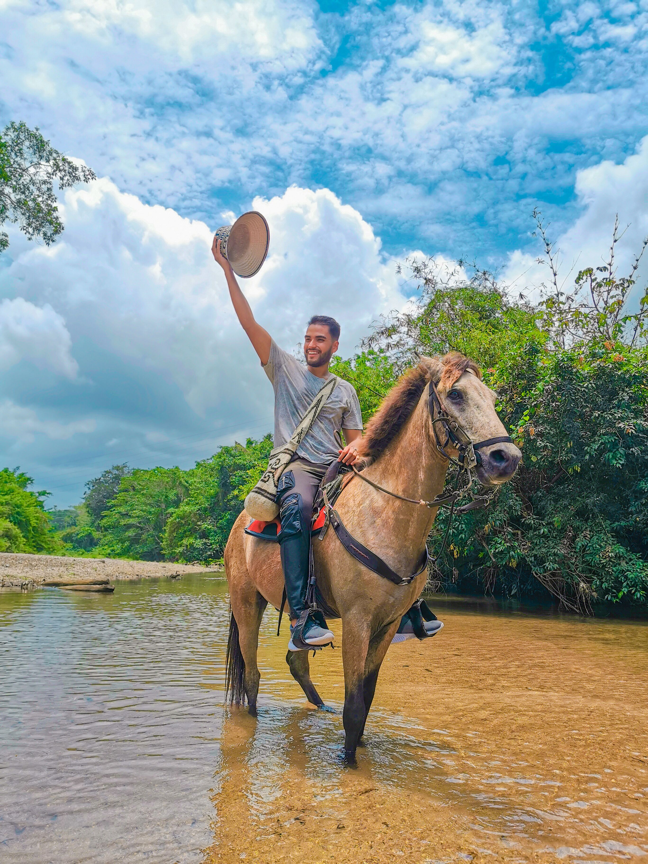 A person riding a horse through a shallow water body, surrounded by lush greenery and a partly cloudy sky.