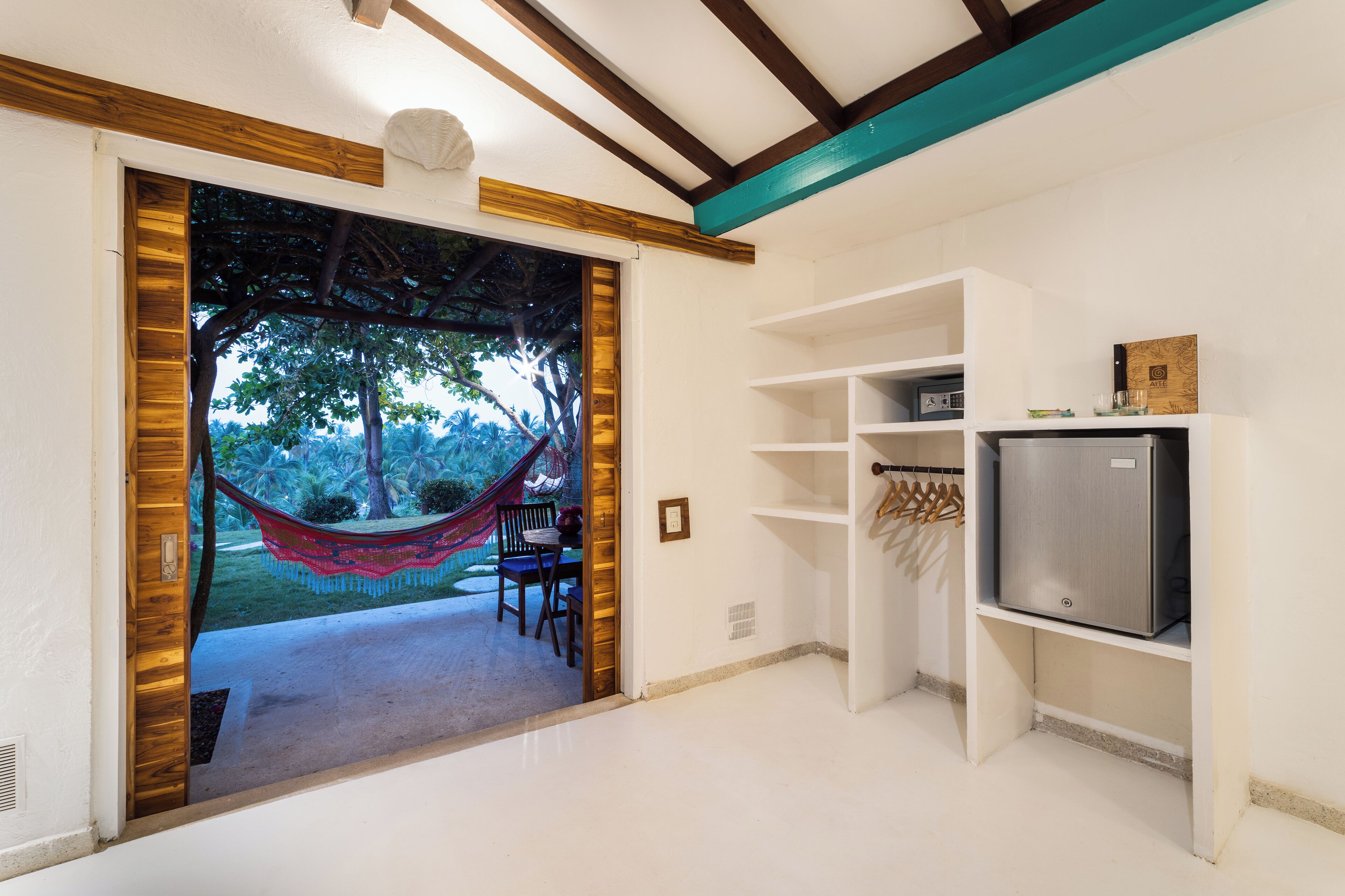 A room with a wooden-framed door leading to a hammock and chairs outside, a white wall with a shelf holding a refrigerator and a small item, and a ceiling with exposed beams.