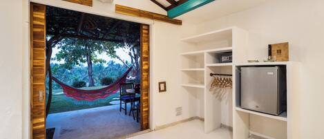 A room with a wooden-framed door leading to a hammock and chairs outside, a white wall with a shelf holding a refrigerator and a small item, and a ceiling with exposed beams.