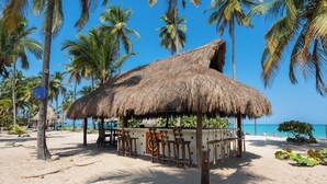 A beach with a thatched-roof hut, palm trees, and a clear blue sky. - Aite Hotel (Dibulla)