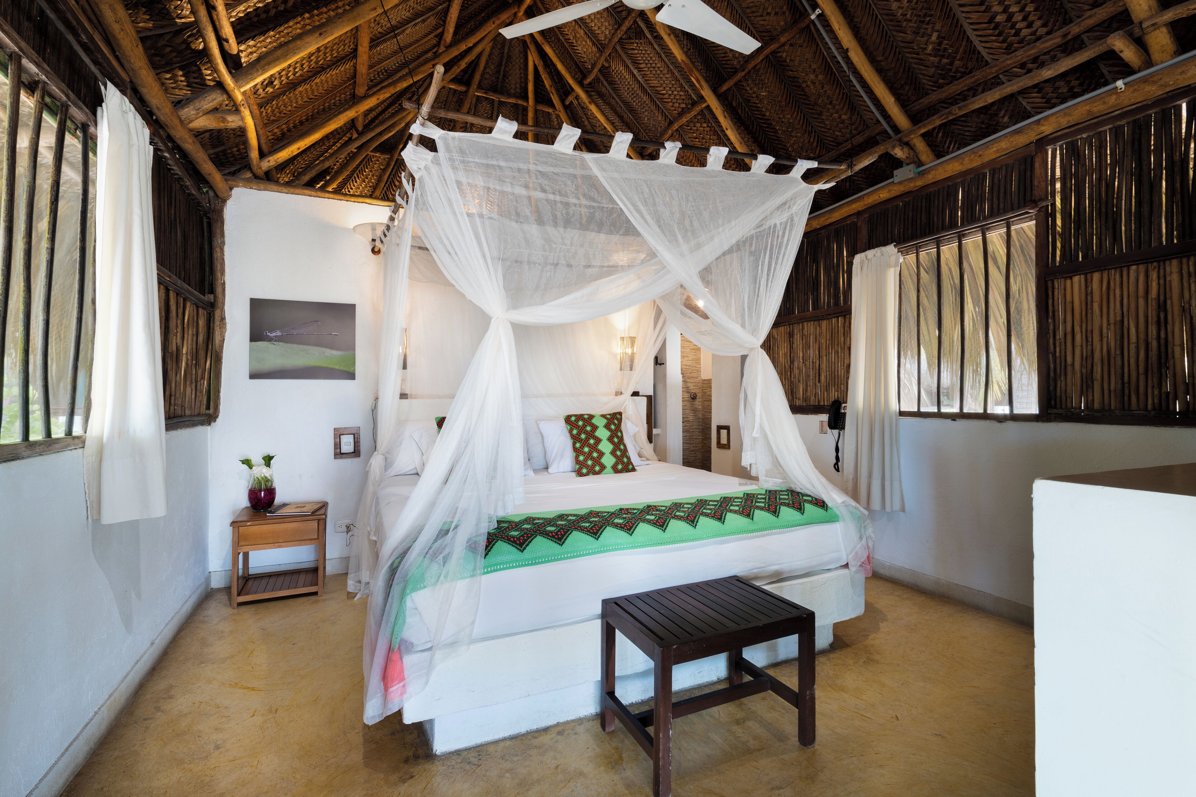 A room with a bed under a canopy, a wooden nightstand with a vase, and a small table.