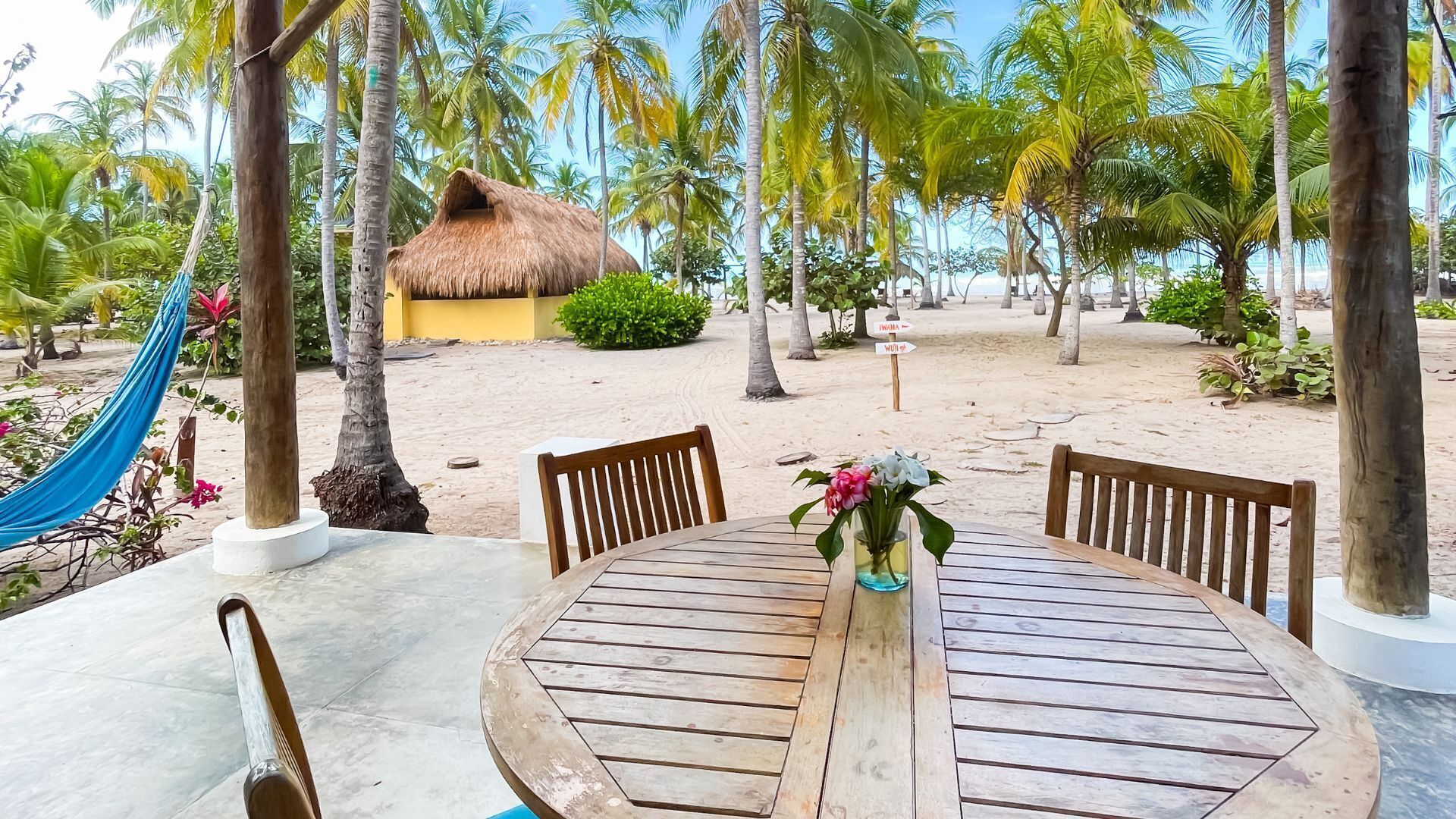 A wooden table with chairs and a vase of flowers, surrounded by palm trees and a thatched-roof structure.