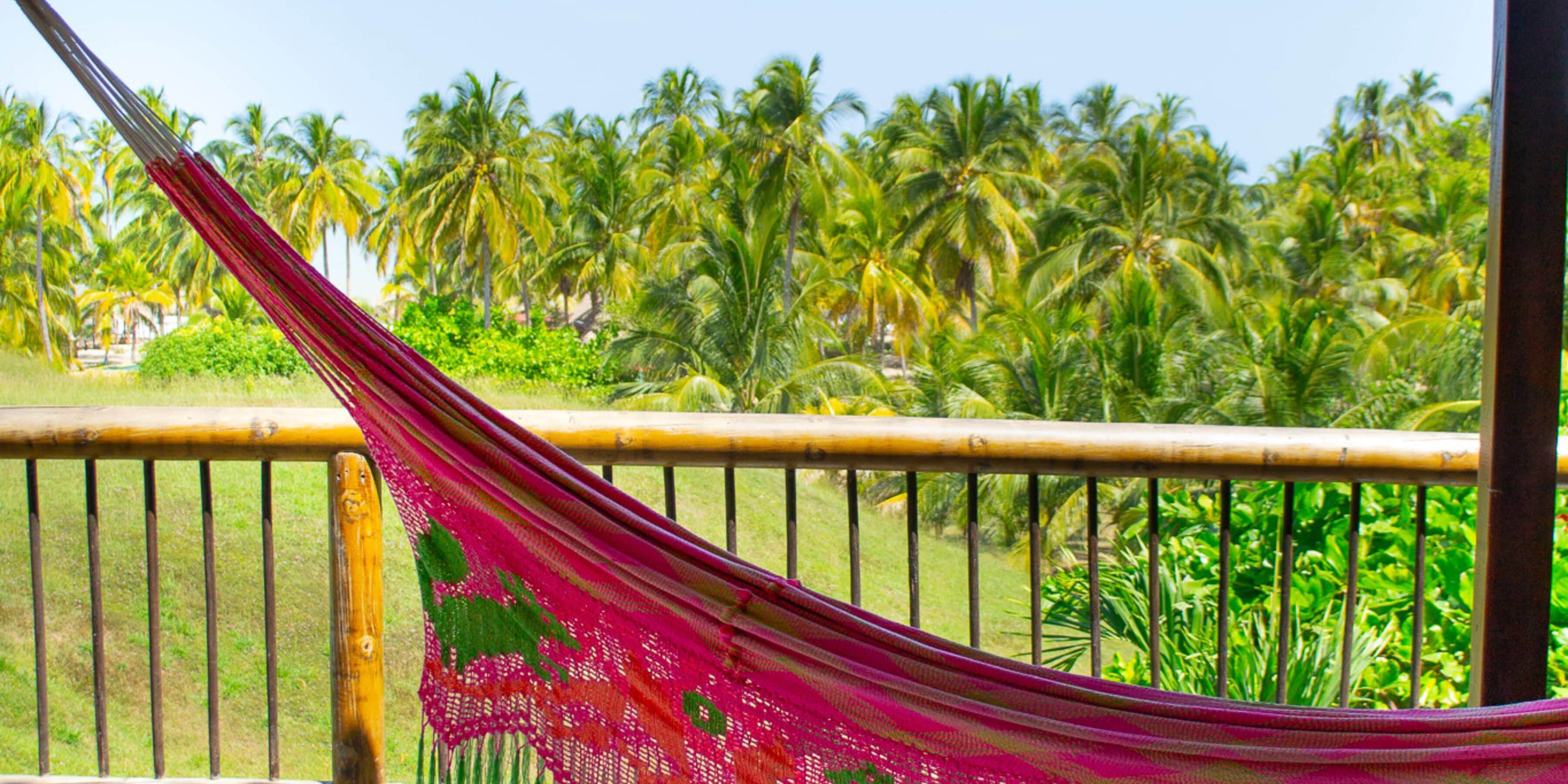 A hammock tied to a wooden railing with a view of palm trees and greenery in the background.