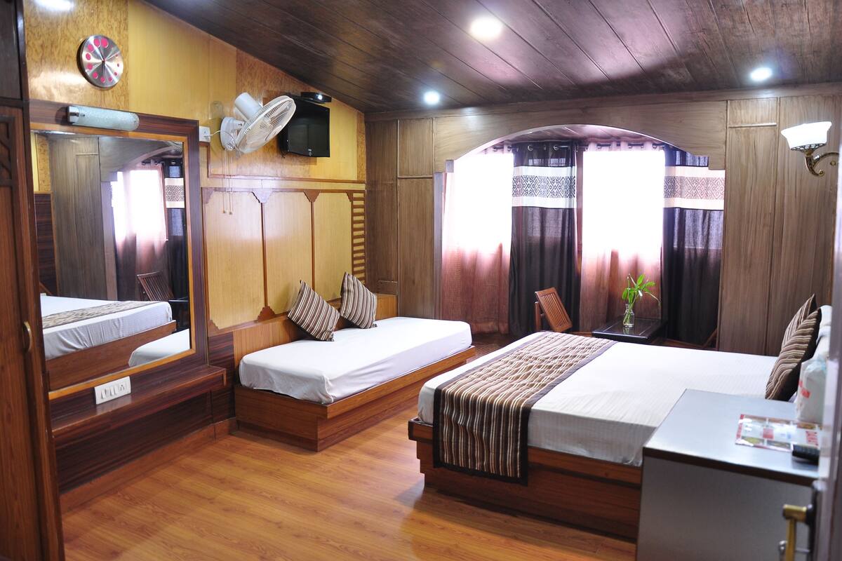 A hotel room with two beds, wooden flooring, a TV, a fan, a desk, and a window with curtains.