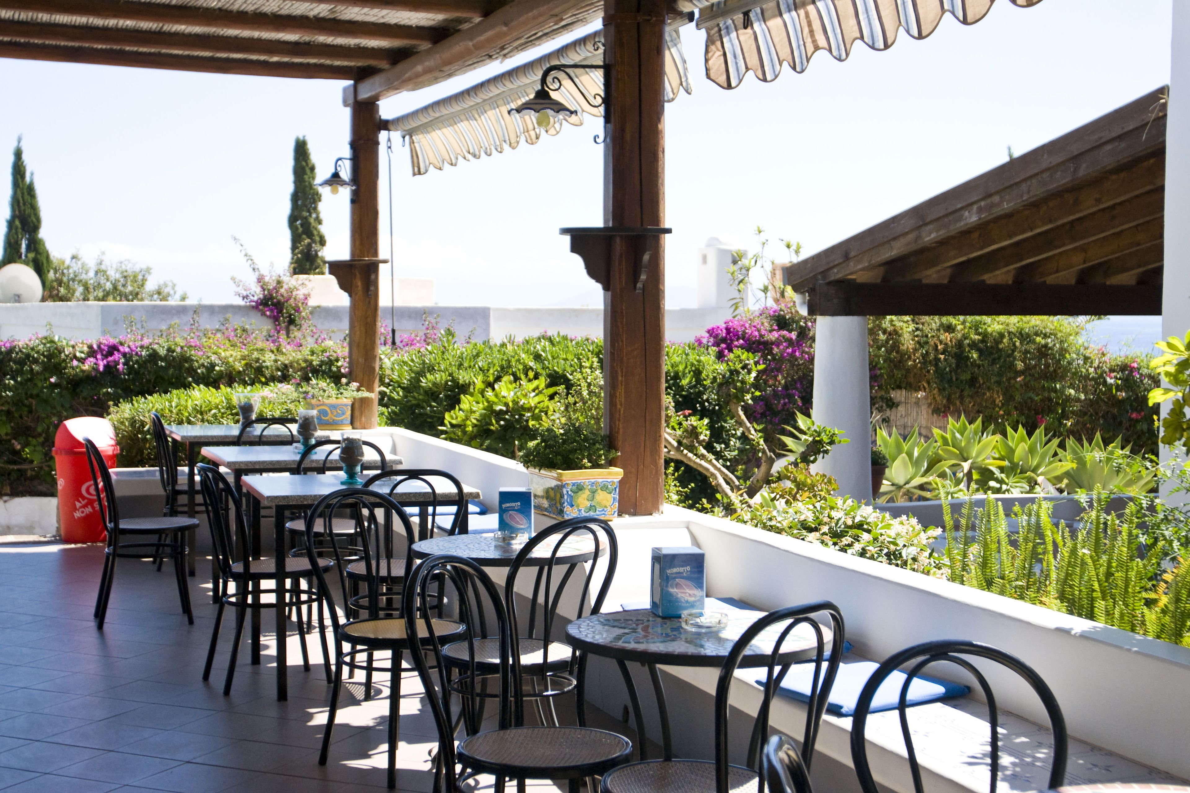 An outdoor patio with tables and chairs, surrounded by lush greenery and a view of the sea.