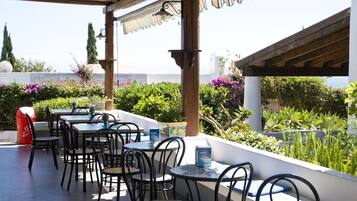 An outdoor patio with tables and chairs, surrounded by lush greenery and a view of the sea.
