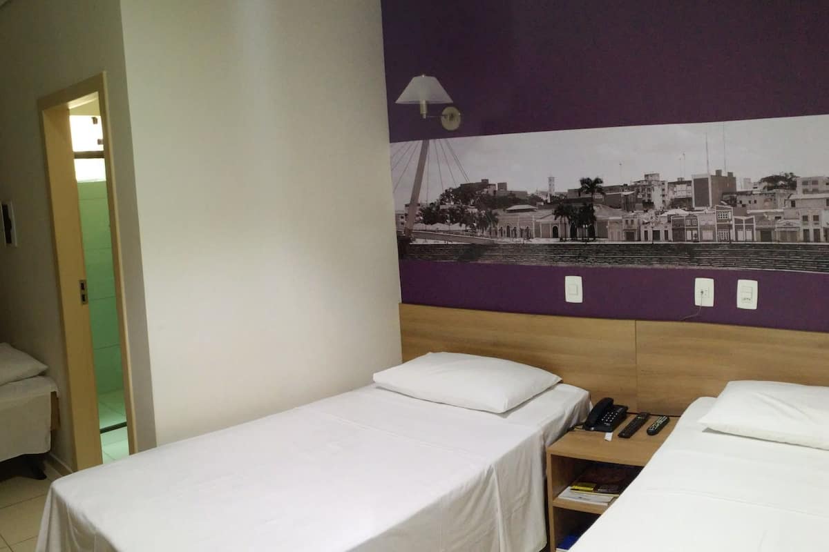 A hotel room with two single beds, a small bedside table with a phone and remote controls, and a wall-mounted lamp.