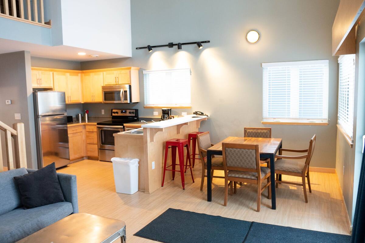 A compact kitchen with modern appliances, a dining area with wooden furniture, and a staircase leading to an upper level.