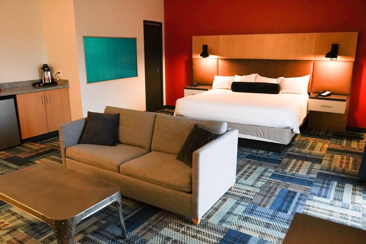 A hotel room with a bed, a sofa, a small table, a nightstand, and a coffee machine.