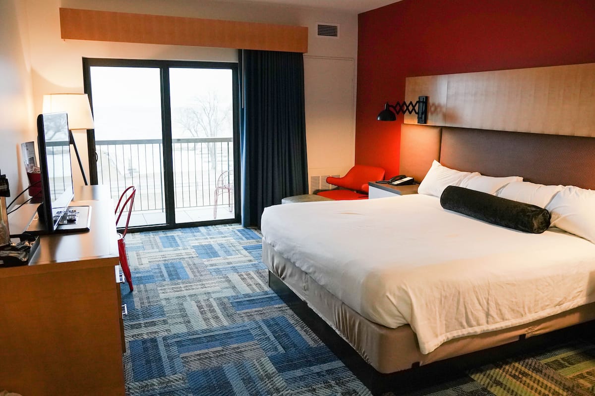 A hotel room with a large bed, a desk with a lamp, a chair, and a view of the outside through a sliding door.