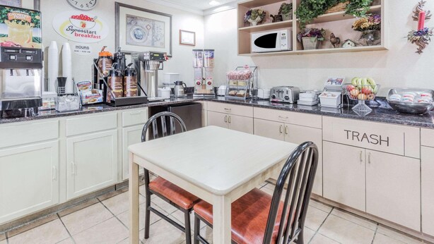A kitchen with a dining table, chairs, a clock, a coffee machine, and a trash bin. - Super 8 by Wyndham St. Charles (SAINT CHARLES)