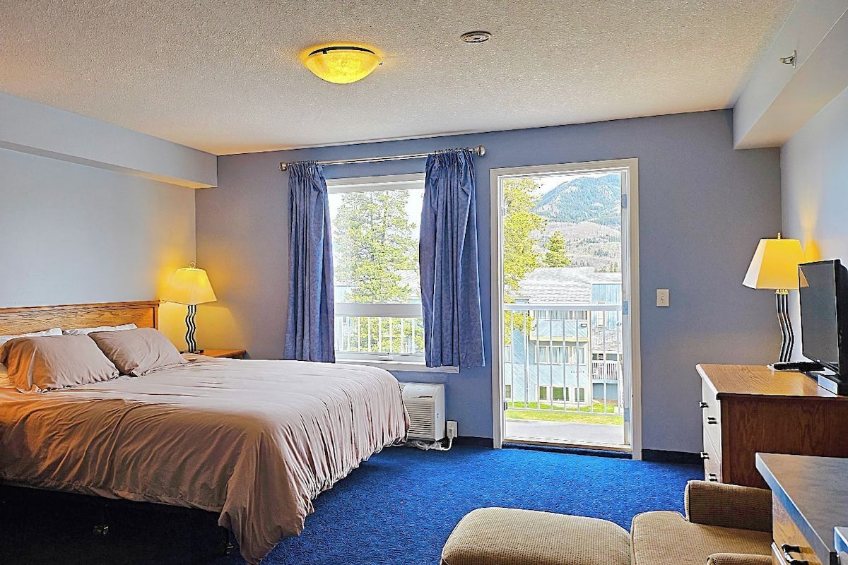 A hotel room with a bed, bedside lamps, a desk, a chair, a television, and a view of the outdoors.