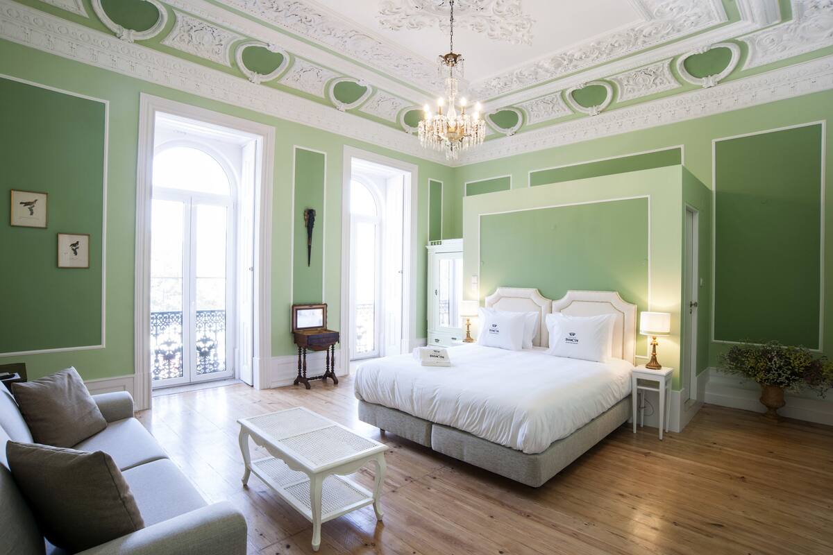 A spacious bedroom with a large bed, a sitting area with a sofa and a small table, and a balcony with a railing.