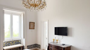 Una habitación con un ventanal grande, una araña, un televisor montado en la pared, un banco y un aparador con espejo.