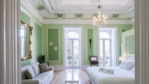 Una habitación con pared verde, techo blanco con molduras decorativas, candelabro, cama, sofá, una mesita y un espejo grande.