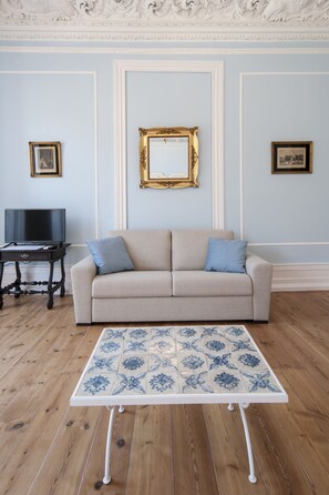 Una sala de estar con un sofá color beige, una mesita con mantel floreado, un televisor sobre un mueble bajo y cuadros enmarcados en la pared.