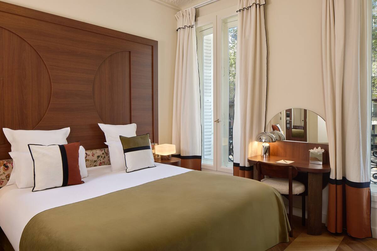 A hotel room with a large bed, a desk with a chair, a mirror, and a window with curtains.